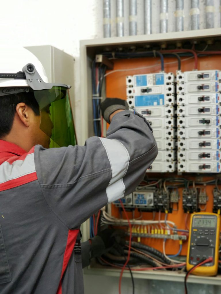 Electrician in a hard hat and face shield testing a panel